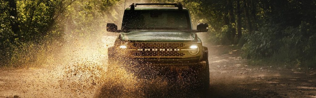 A 2025 Ford Bronco Sport driving through a muddy forest trail near Princeton, IL, with headlights on and dirt splattering into the air.