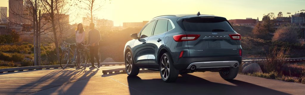 A 2025 Ford Escape parked in a park at sunset near Princeton, IL, with a partial view of downtown and a man and woman walking beside a bicycle.