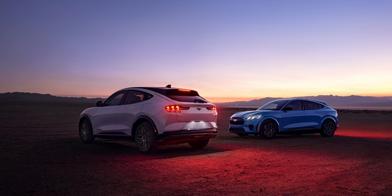 A white and a blue 2025 Mustang Mach-E parked in a desert at sunset near Princeton, IL, with headlights and taillights illuminated.