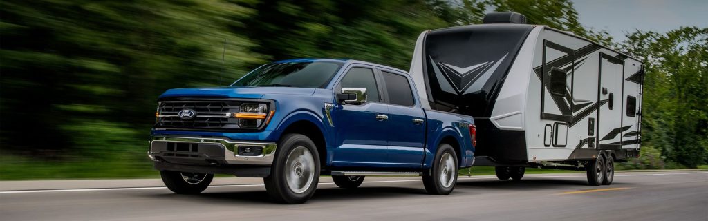 A blue 2025 Ford F-150 towing an RV down a highway surrounded by lush forest near Princeton Ford.