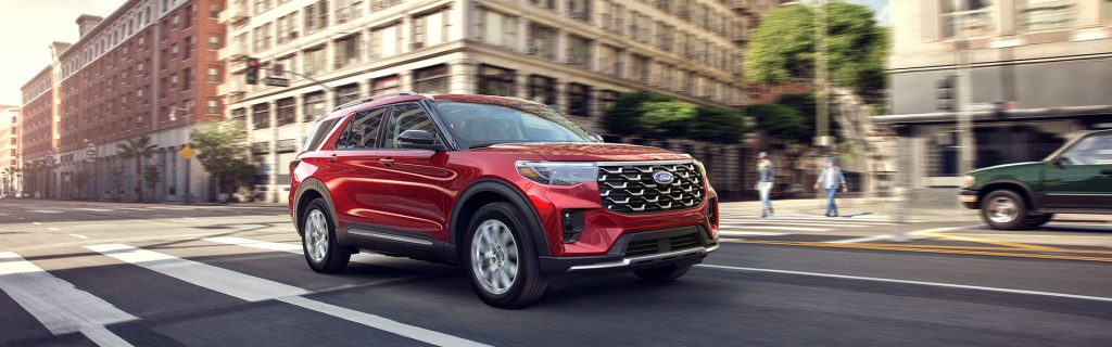 A red 2025 Ford Explorer cruising through busy downtown streets in daylight, surrounded by urban buildings and city life.