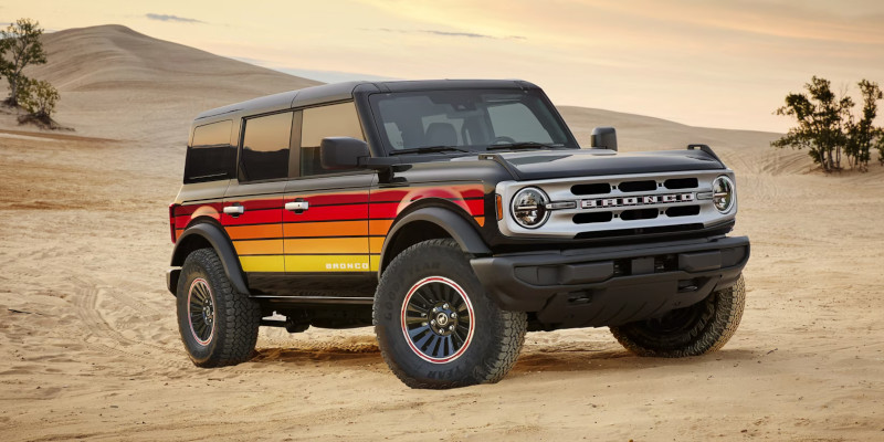 A 2025 Ford Bronco with bold red, orange, and yellow retro-style stripes on the doors driving through a sandy desert under a clear sky.