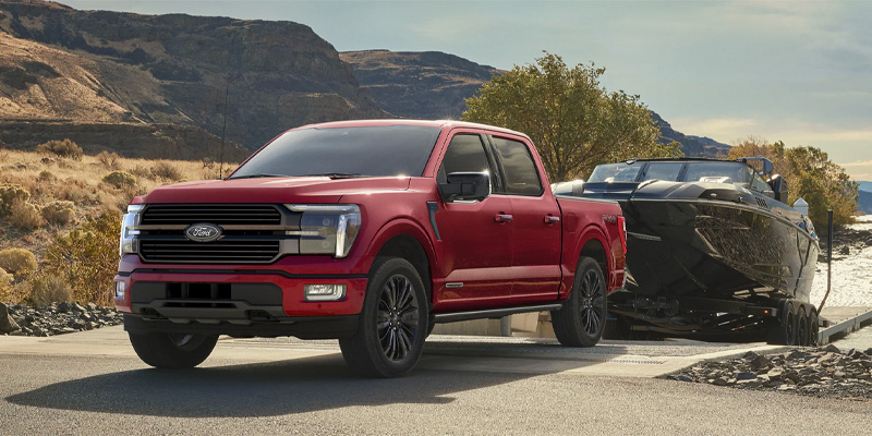 A red 2025 Ford F-150 towing a boat from the water with scenic mountain views in the background near Princeton, IL, highlighting its rugged towing capability.